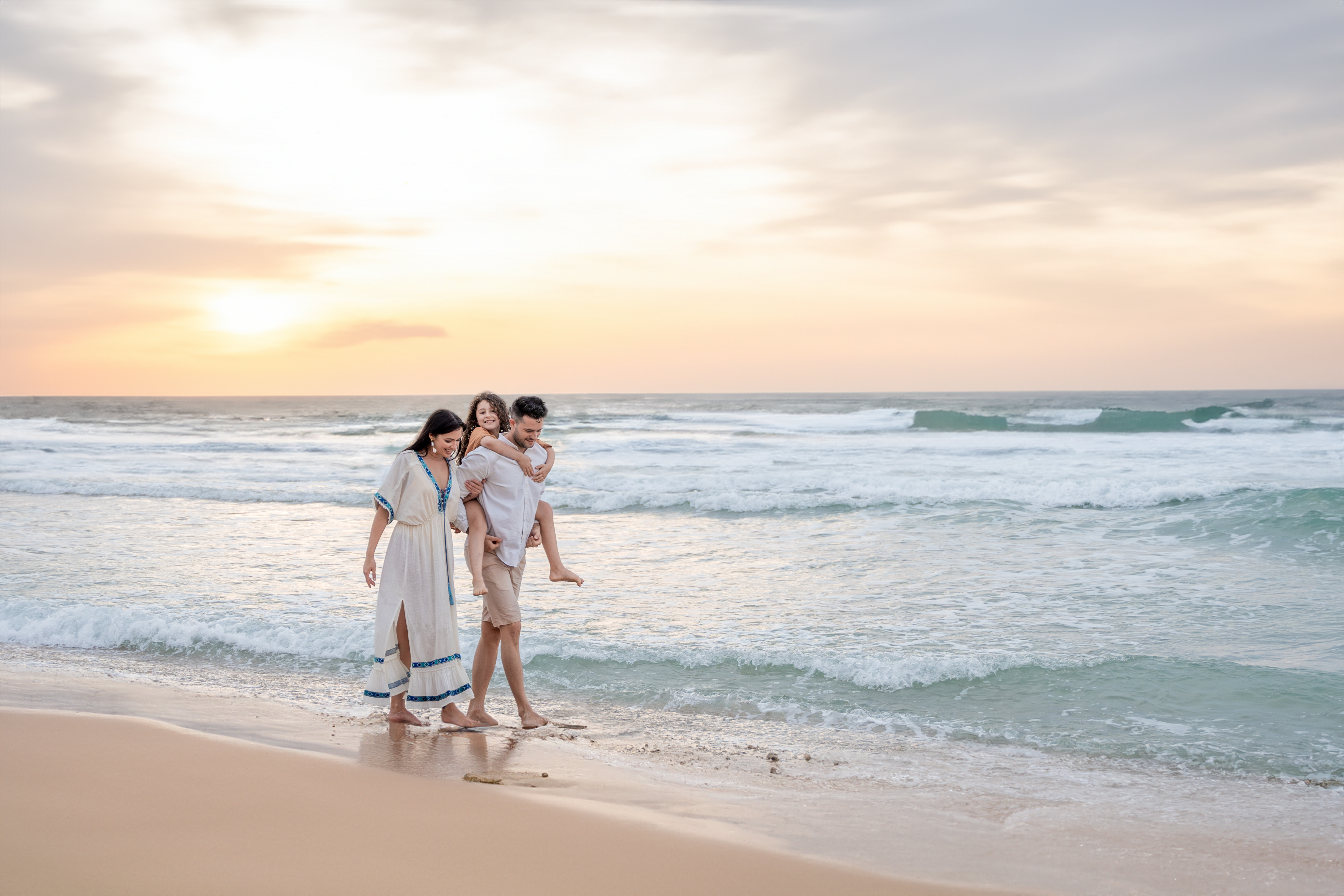 A happy family of three walks barefoot along the shoreline at sunset, with the father giving the child a piggyback ride while the mother walks beside them, all smiling and enjoying the gentle ocean waves and golden sky.